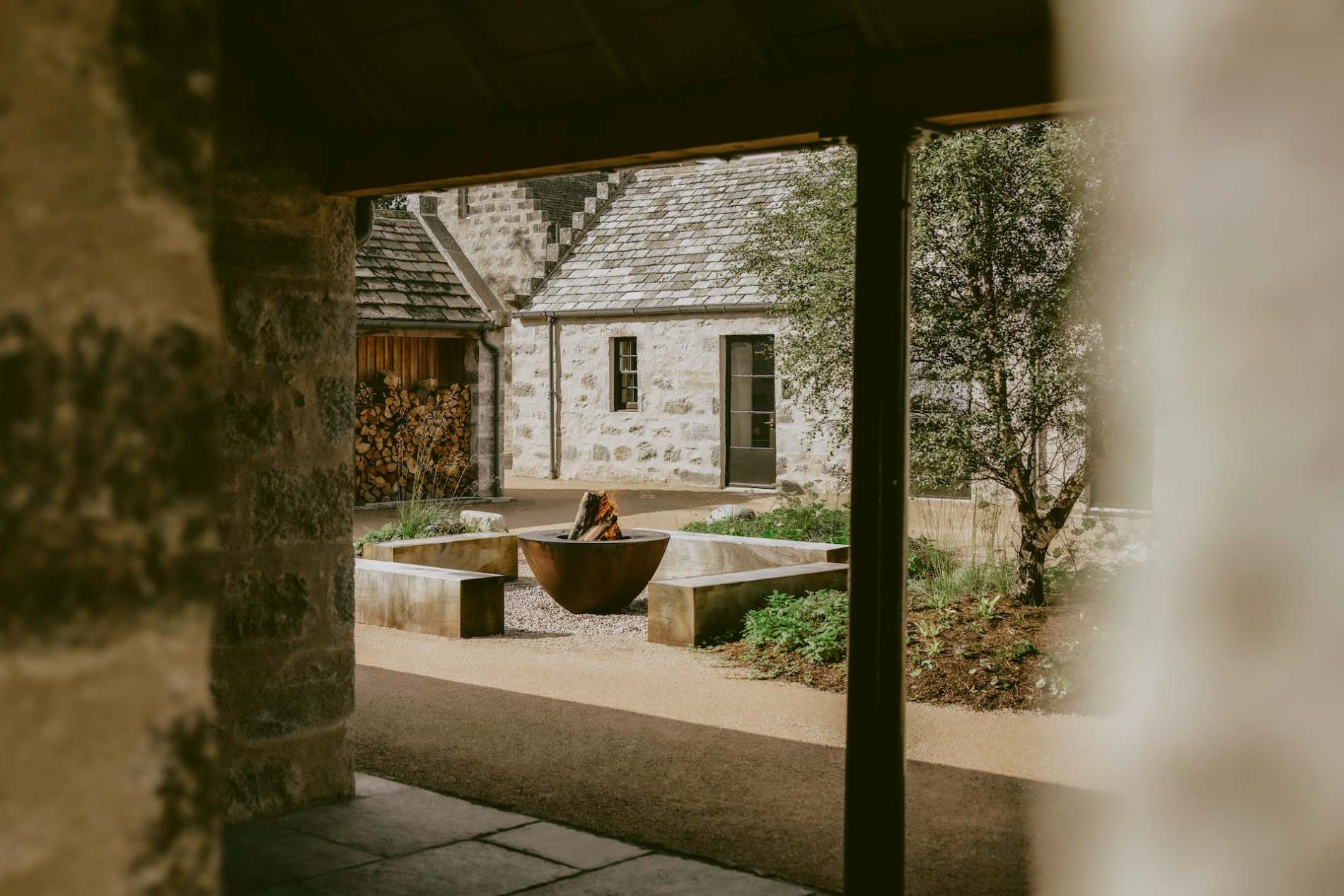 A stone archway frames a courtyard where a fire bowl sits on a gravel bed, stacked logs and a stone farmhouse visible beyond.