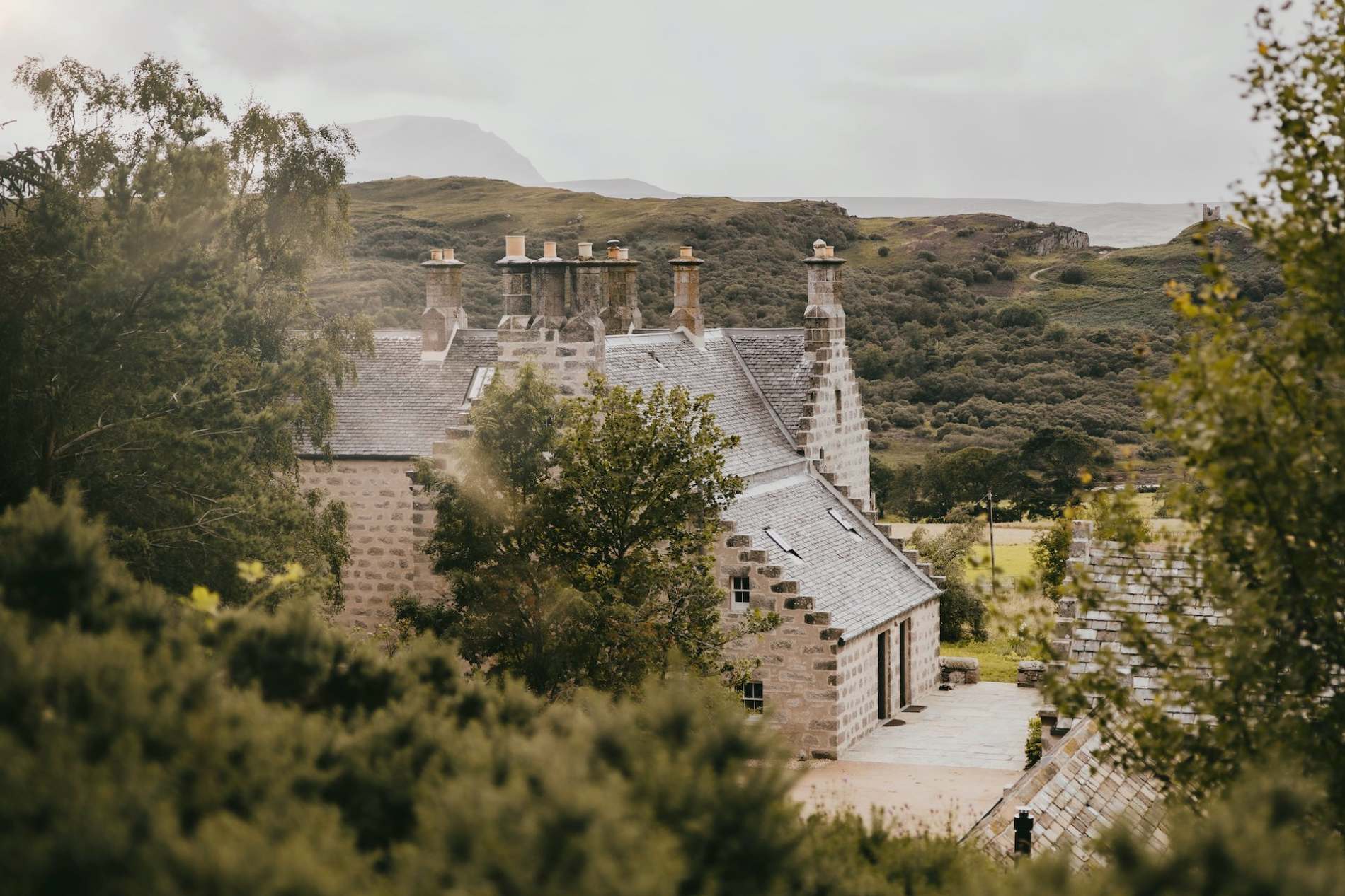 A stone house with crow-stepped gables and clustered chimneys rises above trees, hills visible in the misty distance.