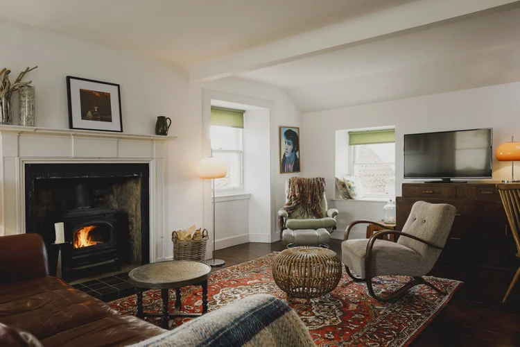A living room has a lit wood burner in a brick fireplace, a leather sofa and a wicker chair on a patterned rug, with a television on a wooden dresser and a framed print above the mantel.