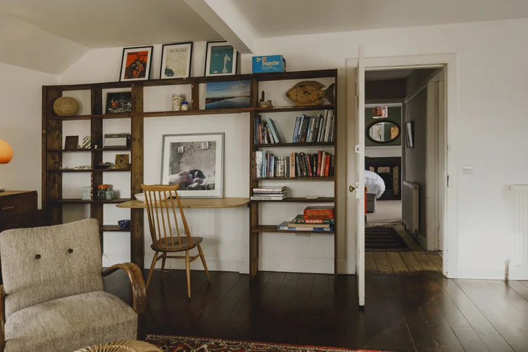 A wooden shelving unit doubles as a desk with a wicker chair, holding books, shells and a framed black-and-white photograph, an armchair in the foreground.