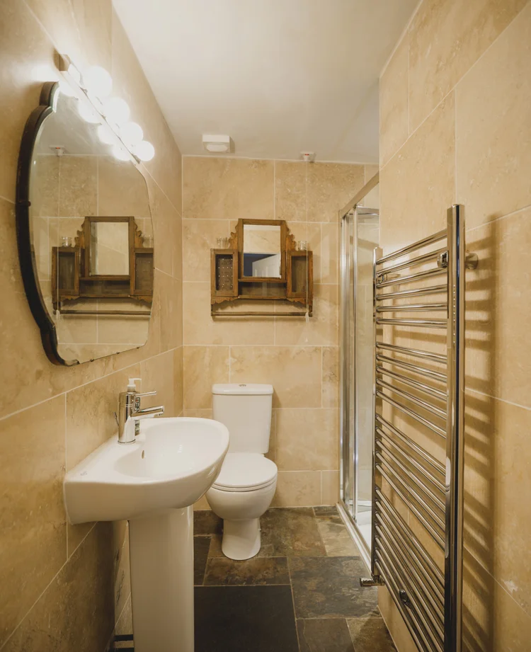 An oval mirror lit by wall lights hangs above a pedestal sink beside a toilet, with a walk-in shower, a chrome heated towel rail and dark tiled flooring.