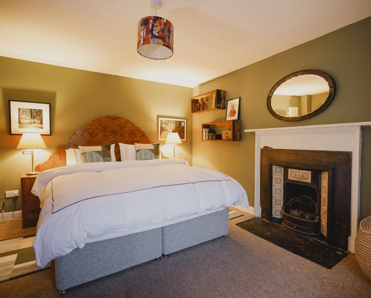 An olive-green bedroom has a curved wooden headboard flanked by bedside lamps, an oval mirror above a tiled fireplace and a small framed picture on a shelf.