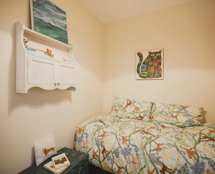 A white wall-mounted shelf unit with a painted landscape and a cat-patterned print hangs above a bed dressed in a monkey-print patterned duvet.