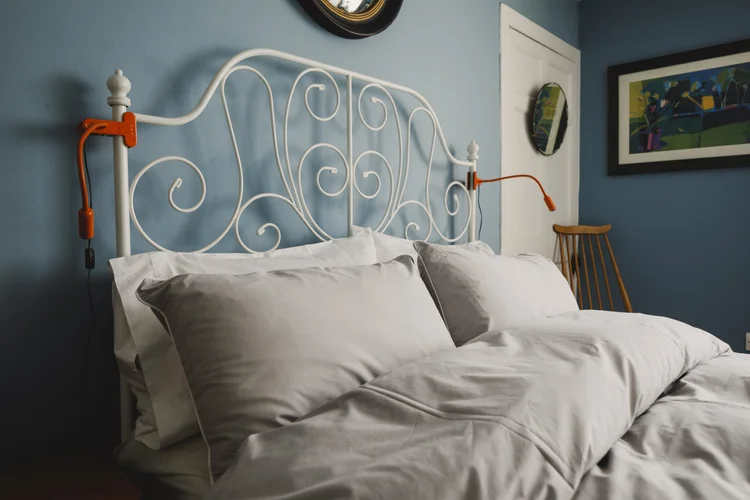 A white wrought iron bed with an ornate scrolled headboard is dressed with grey pillows and a duvet against a blue wall, bedside lamps with orange cords on either side.