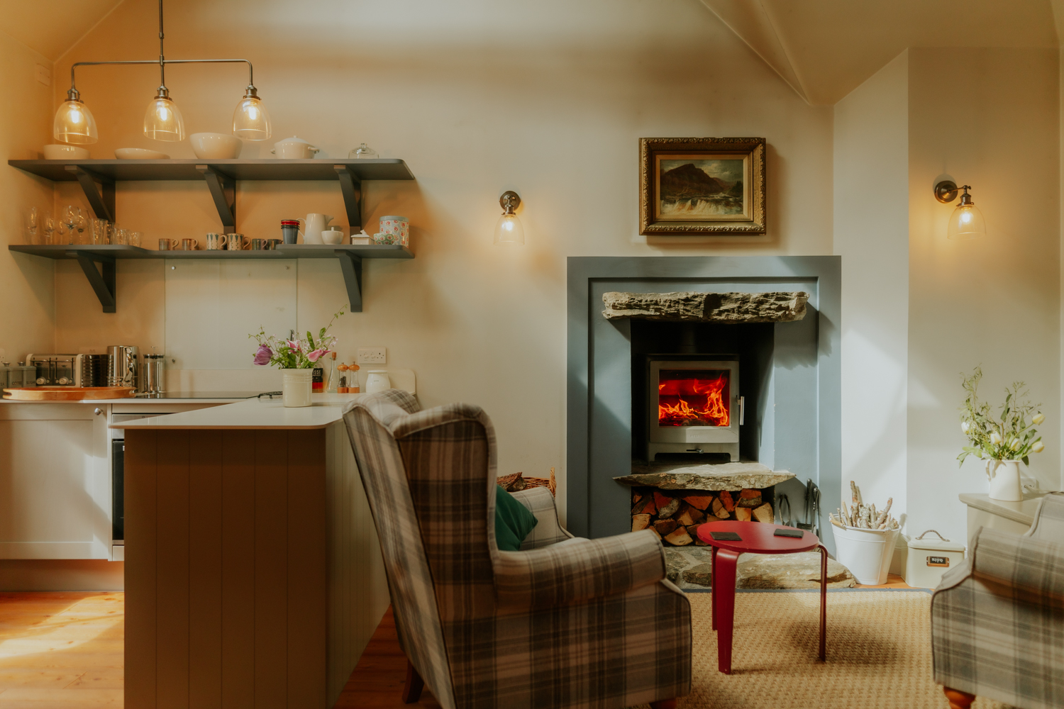 A kitchen with open shelving of crockery and pendant lights stands beside a lit fireplace and two checked armchairs, a red stool and white urns nearby.