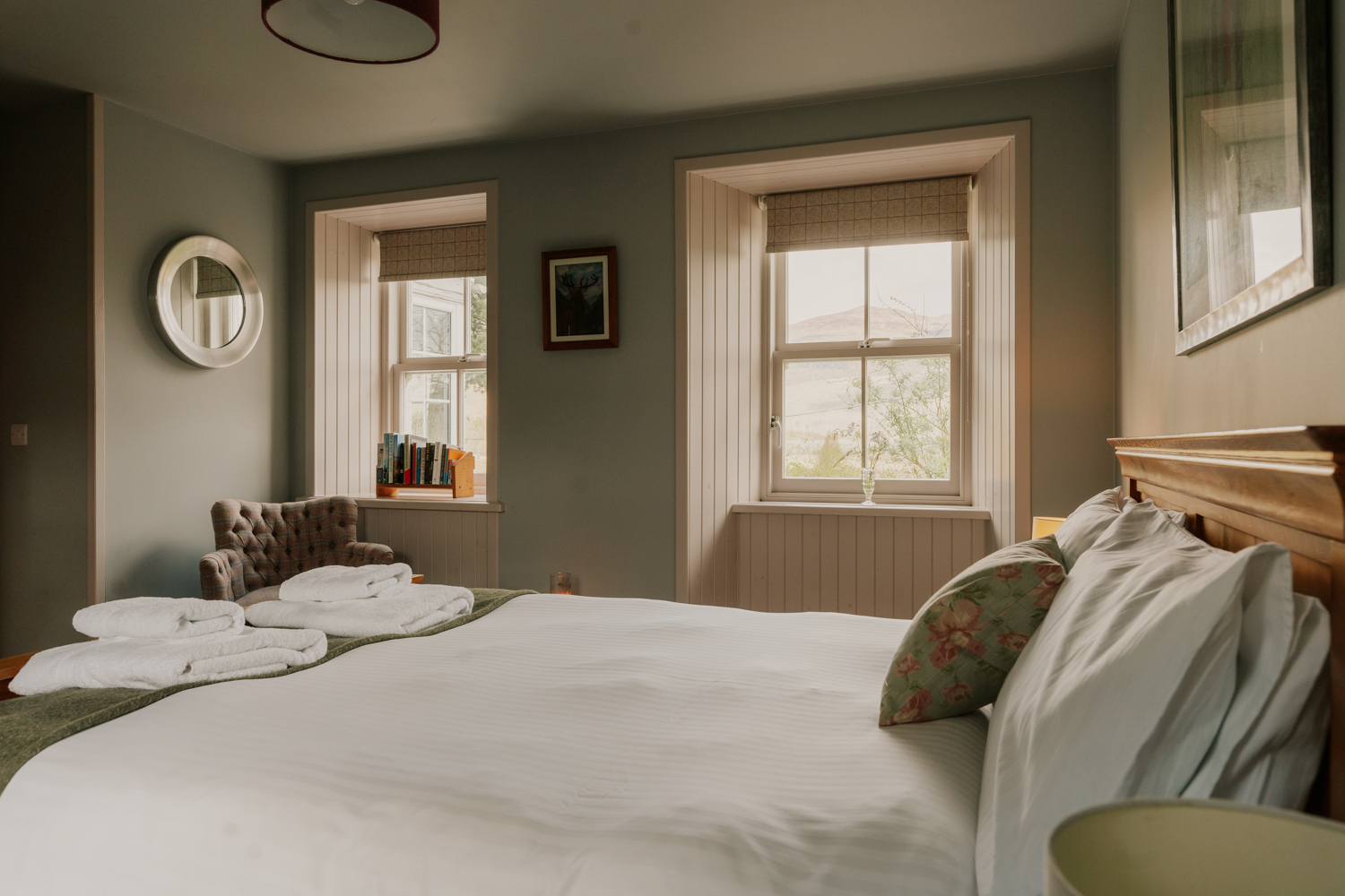 A bed dressed with a floral cushion sits before two curtained windows, towels laid at the foot and a wing-back chair in the corner.