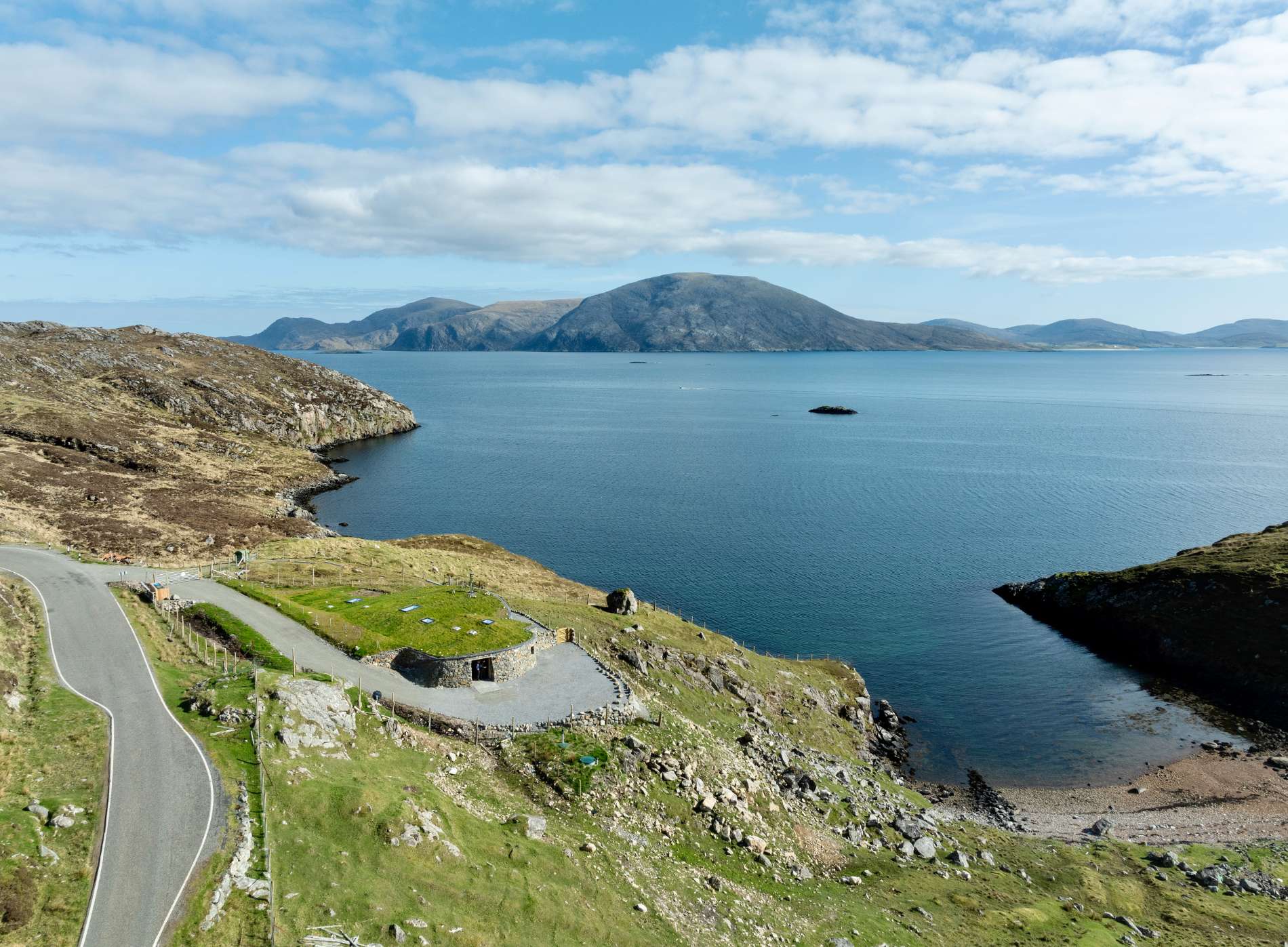 A sea loch stretches between hills, a curved building and jetty visible on the shore below a winding road.