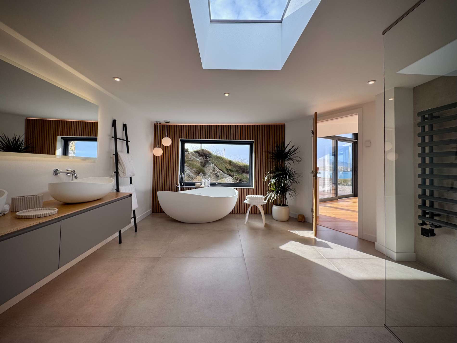 A curved freestanding bath sits beneath a skylight before a window, a vanity with basin to the left and a heated towel rail to the right.
