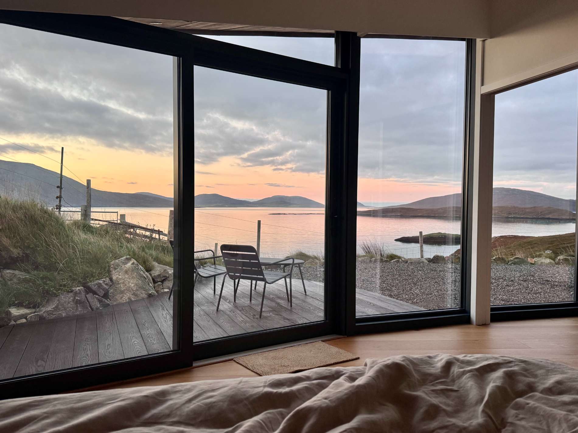 Two chairs stand on a deck beyond floor-to-ceiling windows looking over a beach and hills at sunset.