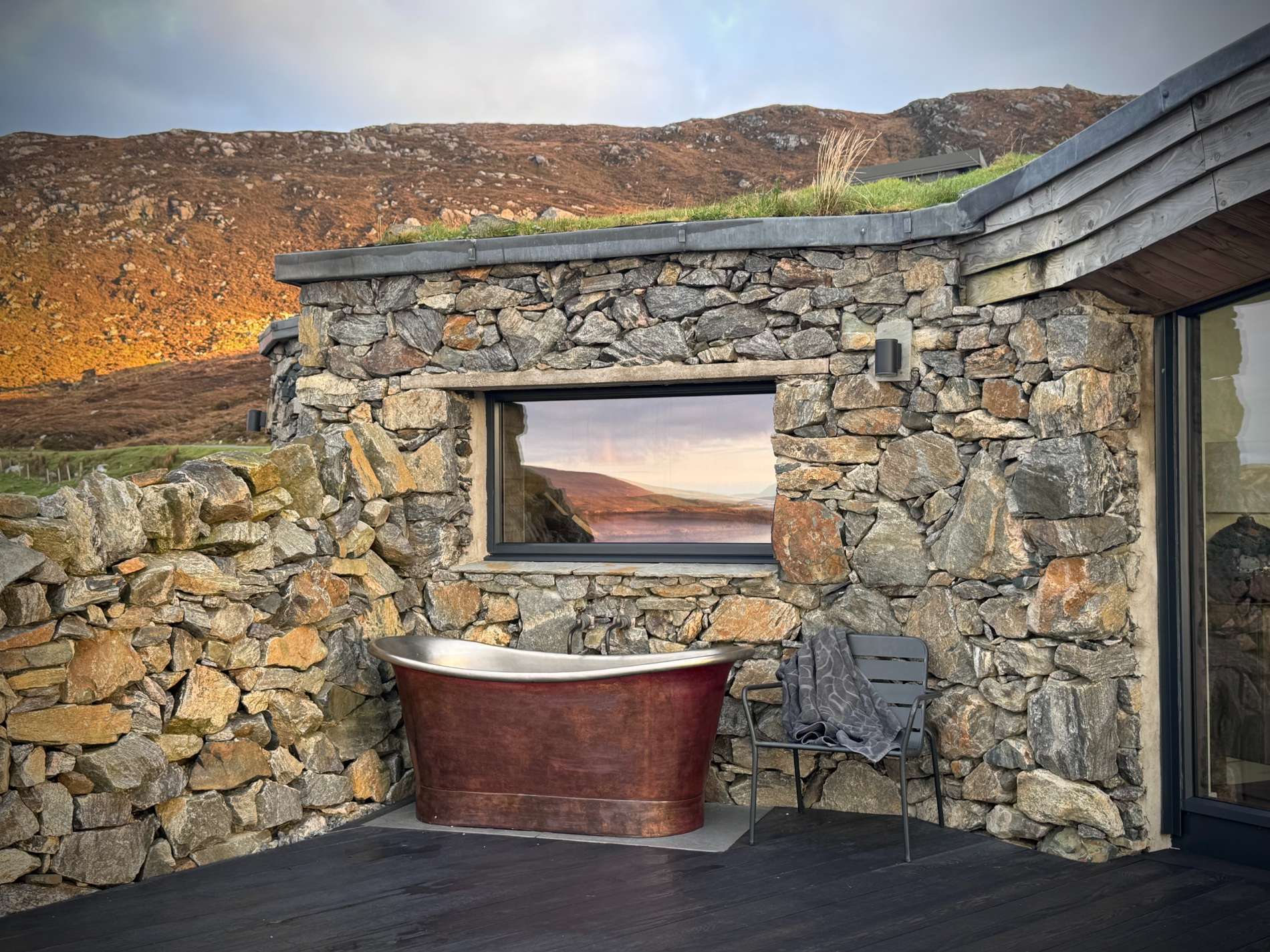 A copper freestanding bath stands against a dry stone wall, a window framing hills beyond and a chair with a towel to the side.