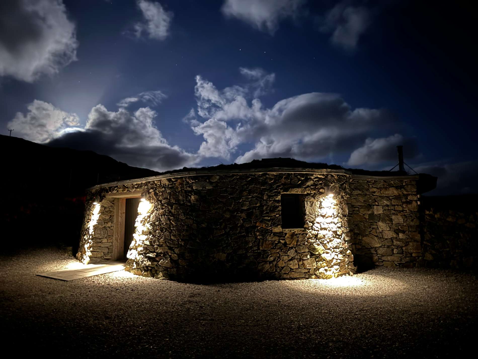 A round stone building glows under a night sky scattered with stars, light spilling from its open door and window.
