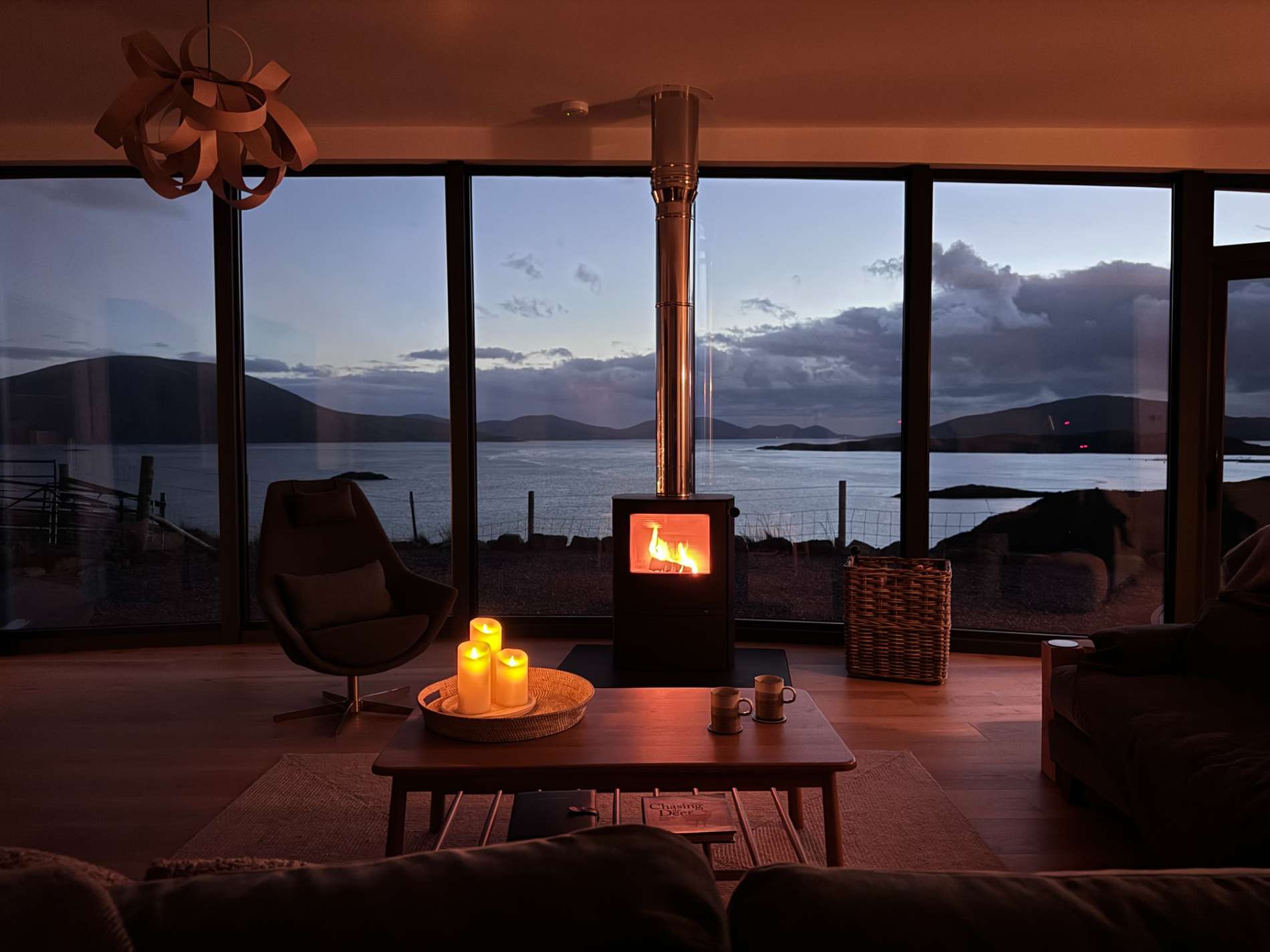 A wood burning stove glows at dusk before floor-to-ceiling windows overlooking a loch, candles lit on the low table in front.