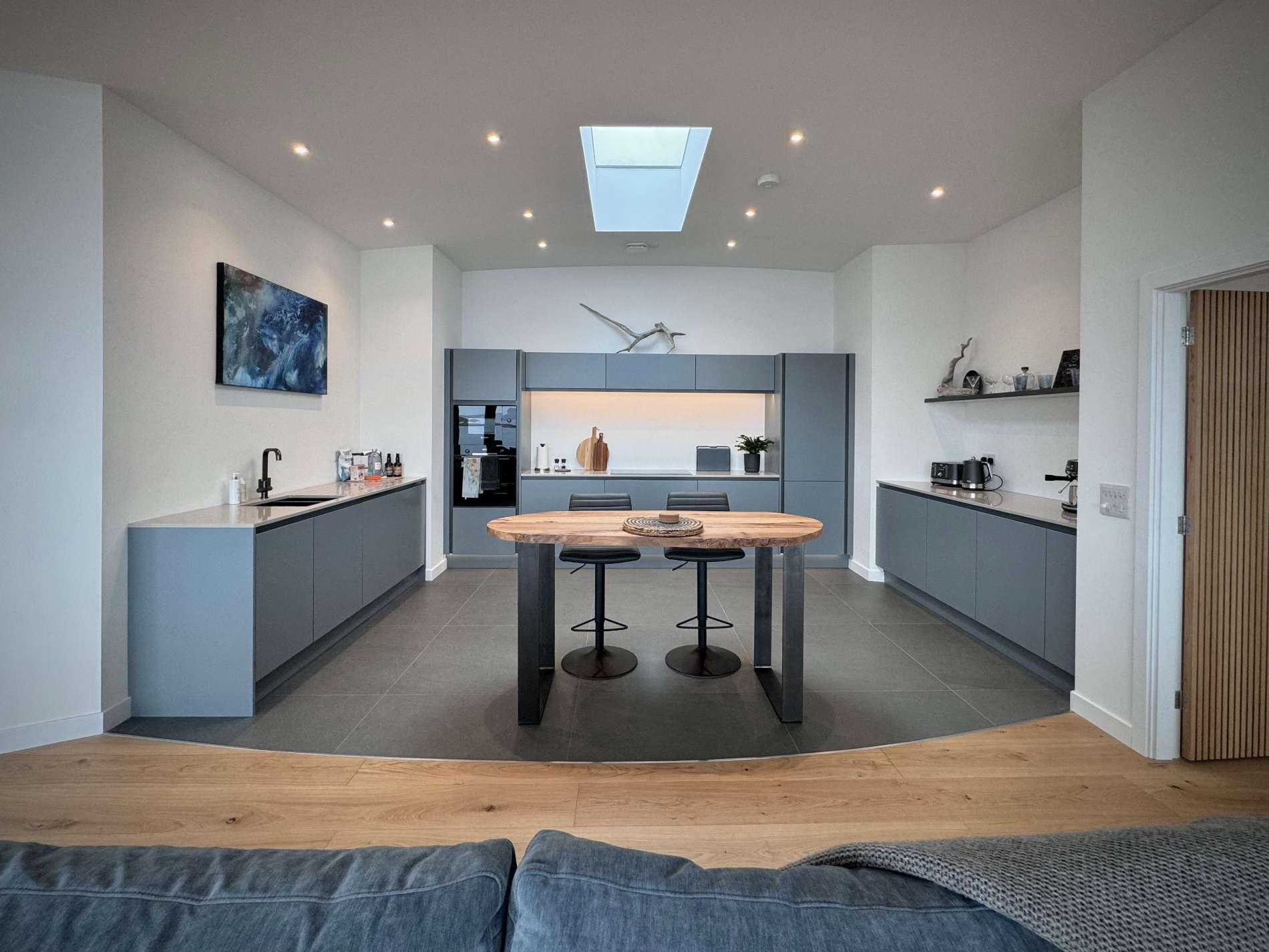 A slate blue kitchen with a central island and bar stools stands below a roof window, a wall-mounted television to the left.