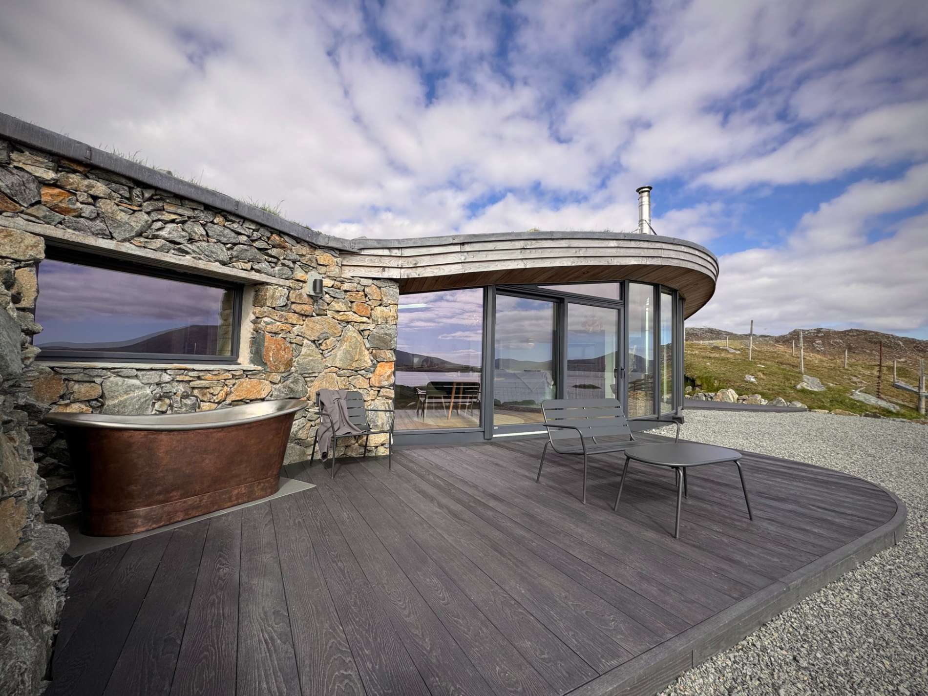 A copper bath stands against a stone wall on a decked terrace, glazed doors and windows curving around the timber-roofed house behind.