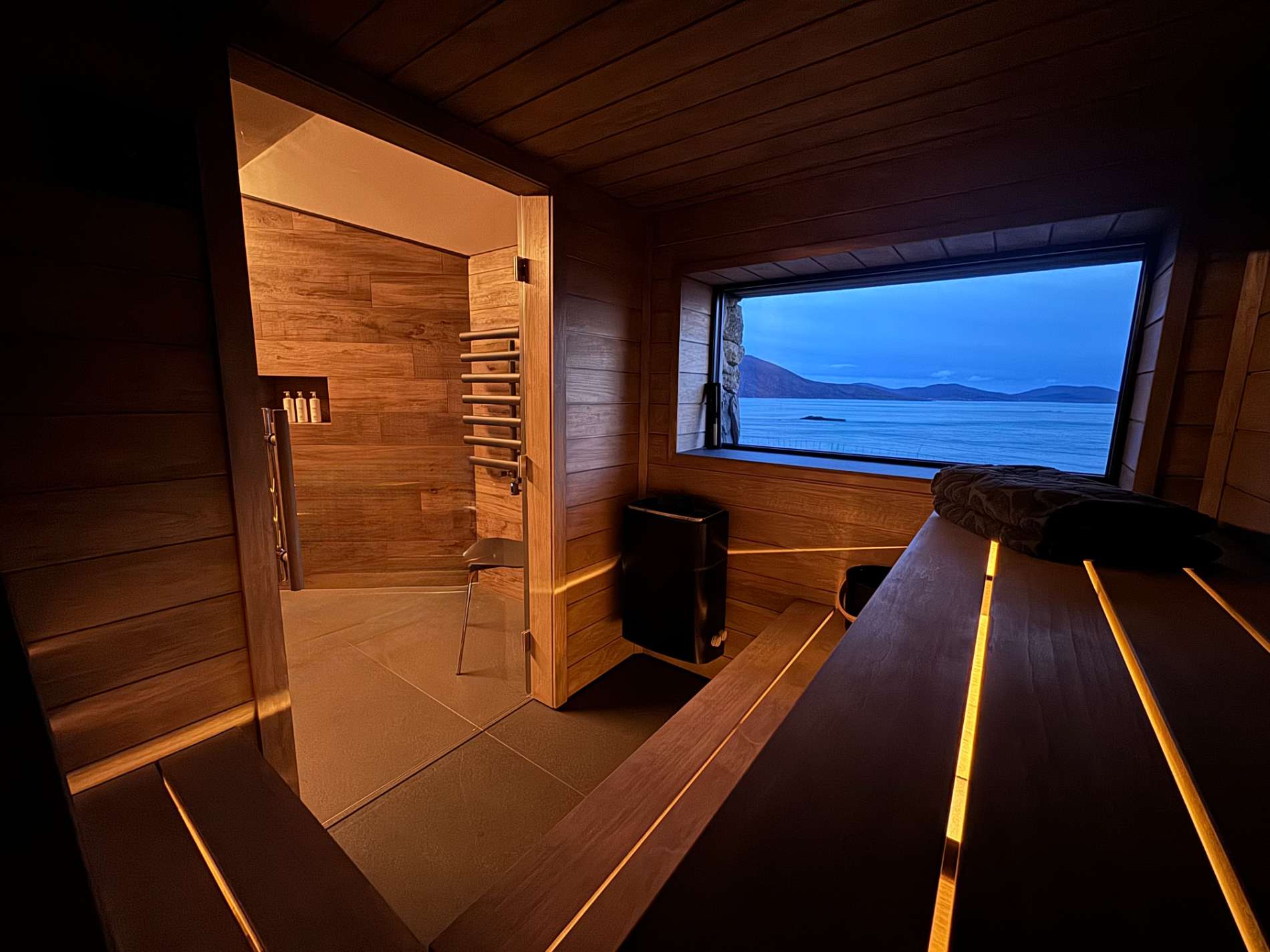 A sauna's glass door opens onto a shower room, a heater and bucket in the foreground beneath a window looking onto the sea at dusk.