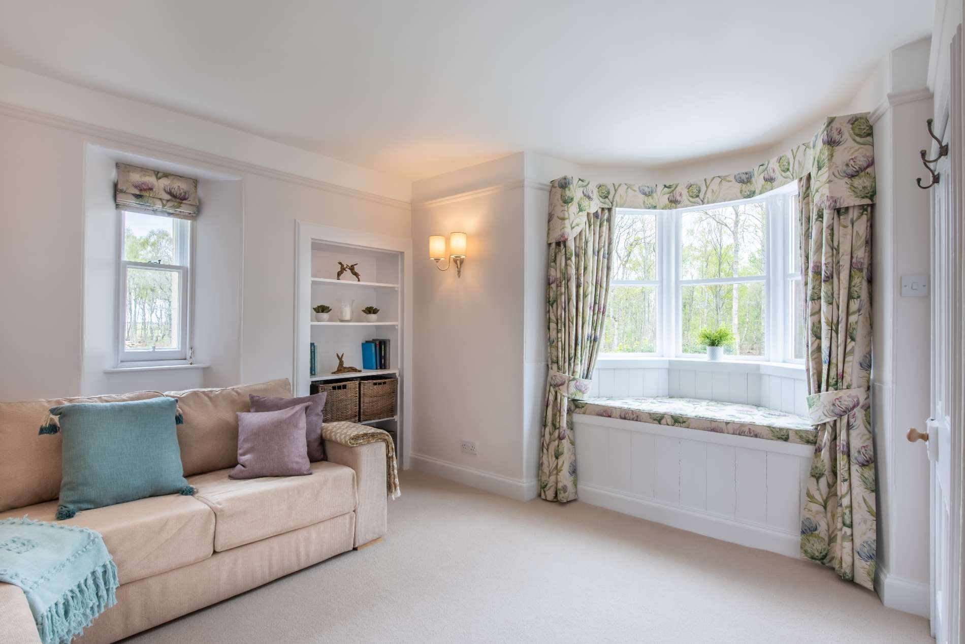 A cream sofa with teal and lilac cushions sits opposite a wall-mounted bookshelf, a bay window seat dressed with floral curtains beyond.