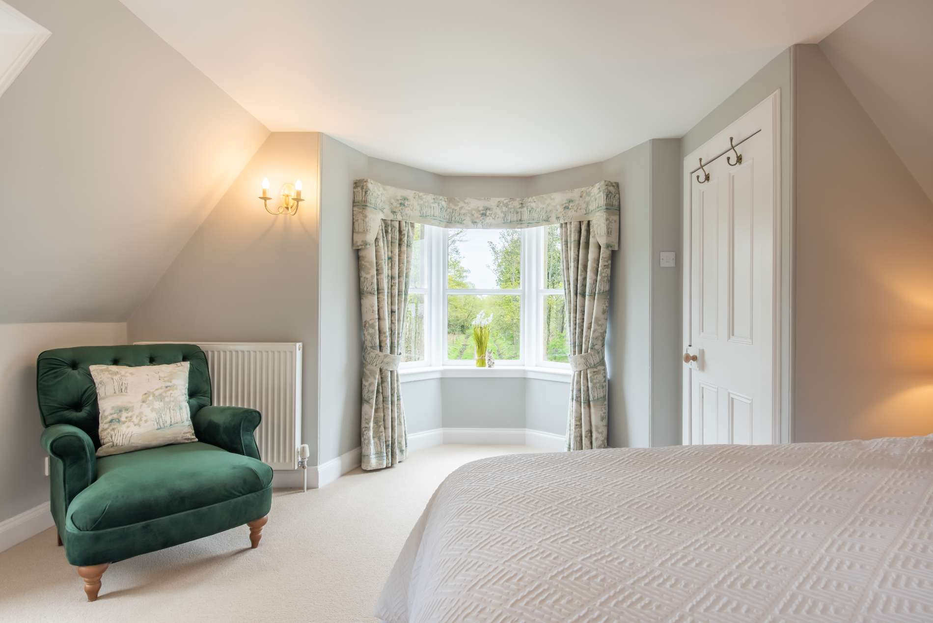 A green velvet armchair with a floral cushion stands in the corner of a bedroom beside a bay window with floral curtains.