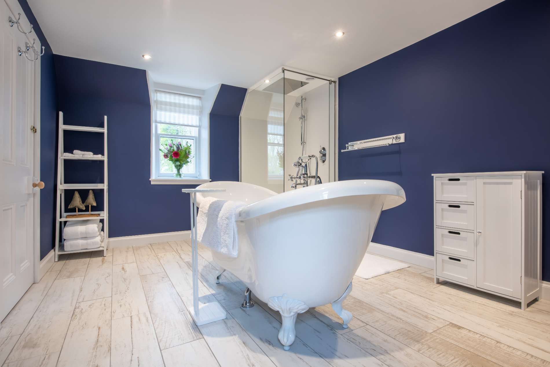 A freestanding roll-top bath stands against deep navy walls beside a glass shower enclosure, a ladder towel rail and a white chest of drawers.
