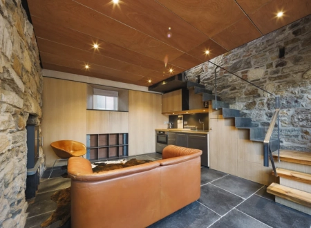 An orange leather sofa draped with a sheepskin faces a stone fireplace beneath a timber ceiling, a staircase rising behind.