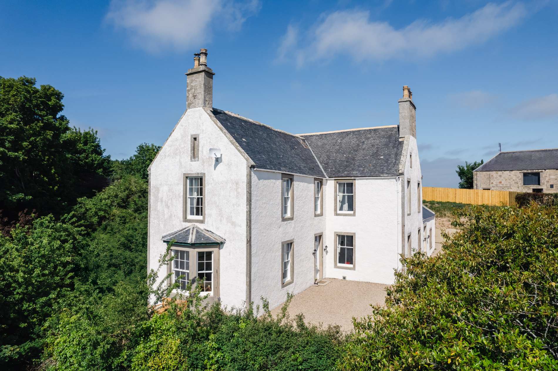 A white harled farmhouse with a slate roof and chimneys stands amid greenery under a blue sky.