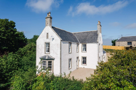 A white harled farmhouse with a slate roof and chimneys stands amid greenery under a blue sky.