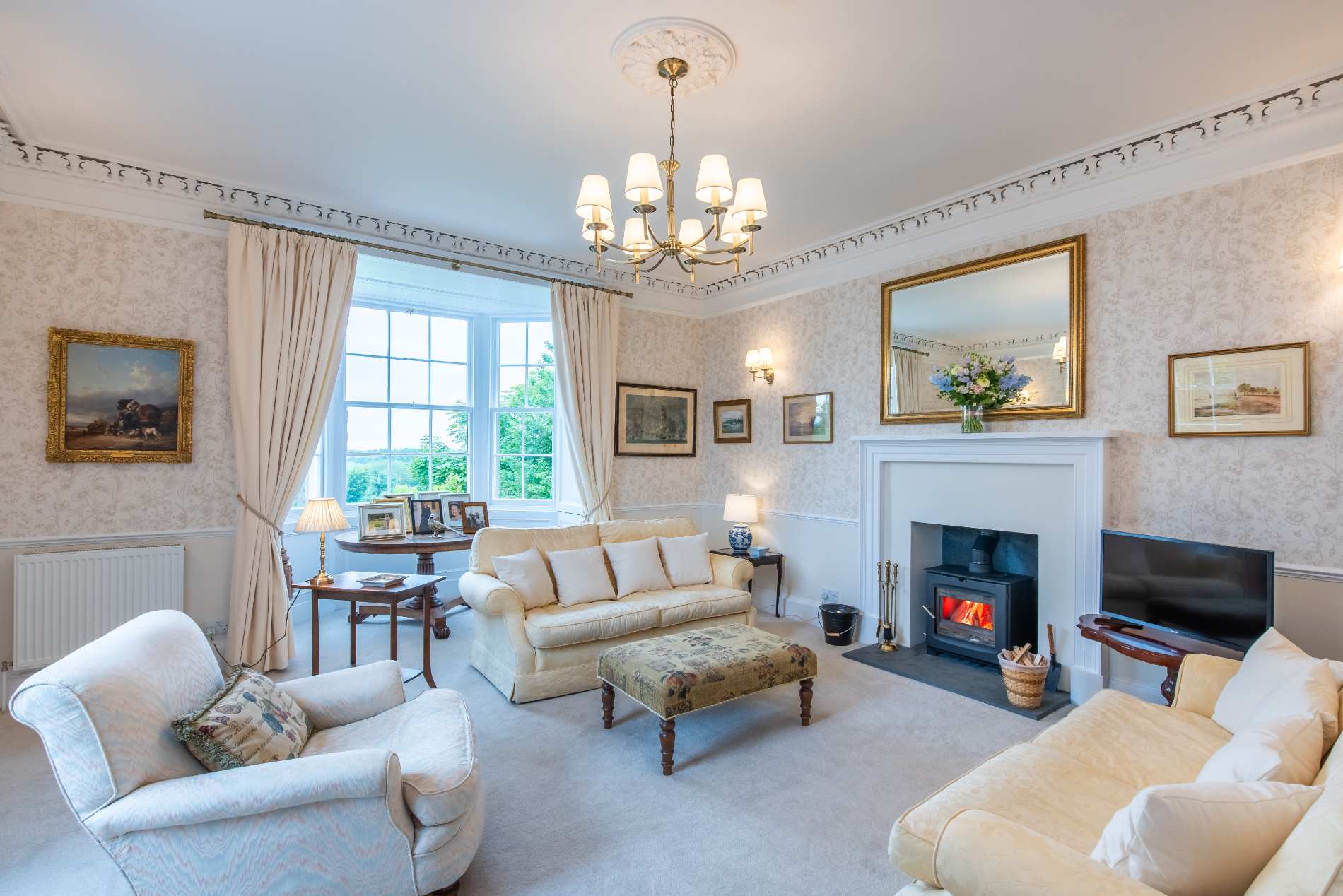 Cream sofas face a wood-burning stove beneath gilt-framed paintings in a sitting room with patterned wallpaper and a chandelier.