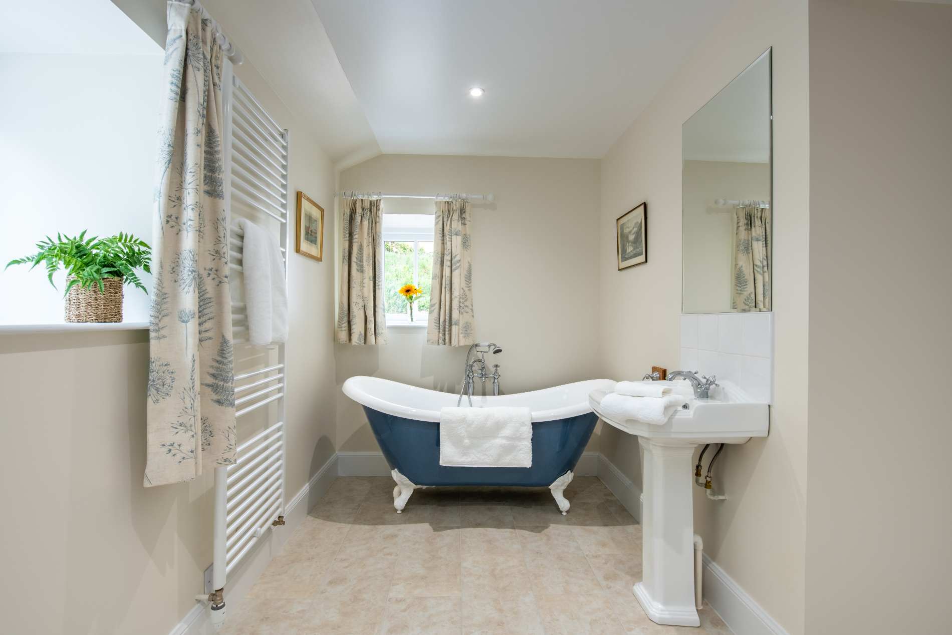 A blue freestanding bath with a white interior stands beside a heated towel rail and pedestal sink, floral curtains at the window.