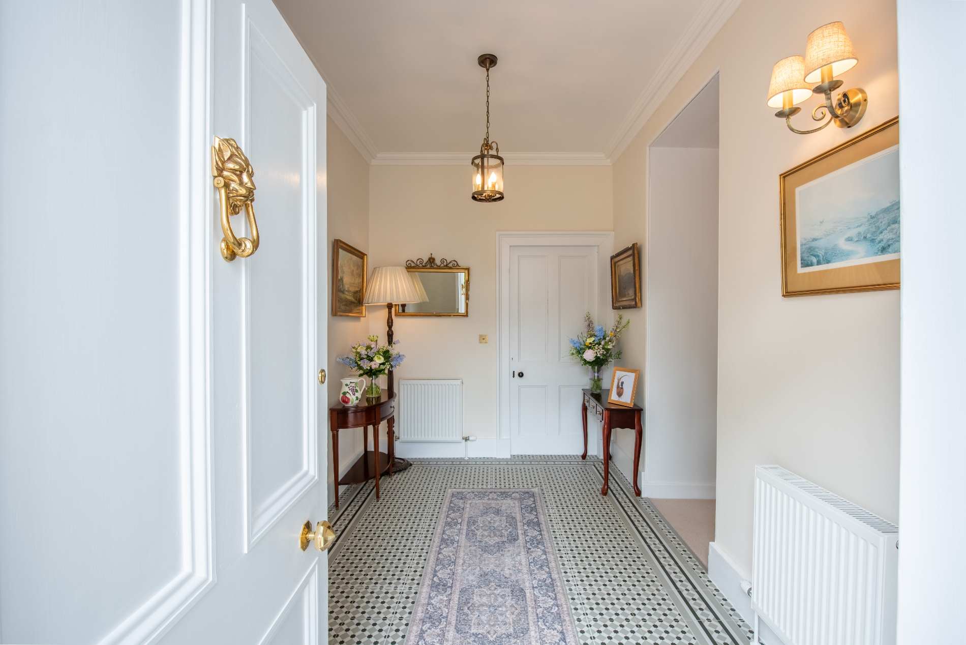 A patterned tiled hallway leads past console tables of flowers to an ornate mirror and framed painting beneath a pendant lantern.