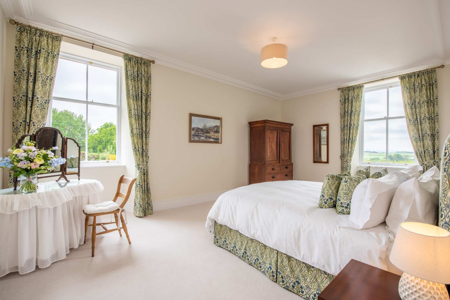 A bedroom with matching floral curtains and bed valance has a dark wood wardrobe and a dressing table set with flowers.