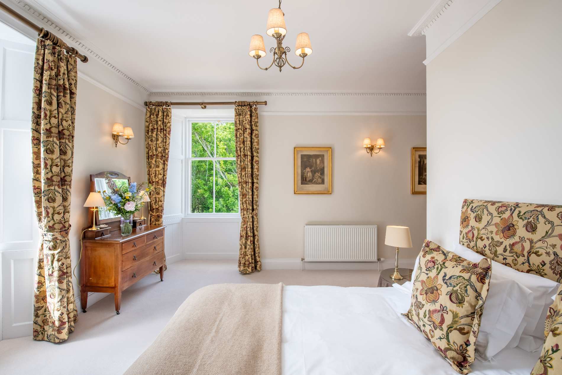 An antique chest of drawers holds a vase of flowers beneath framed pictures, floral curtains and cushions completing the bedroom.