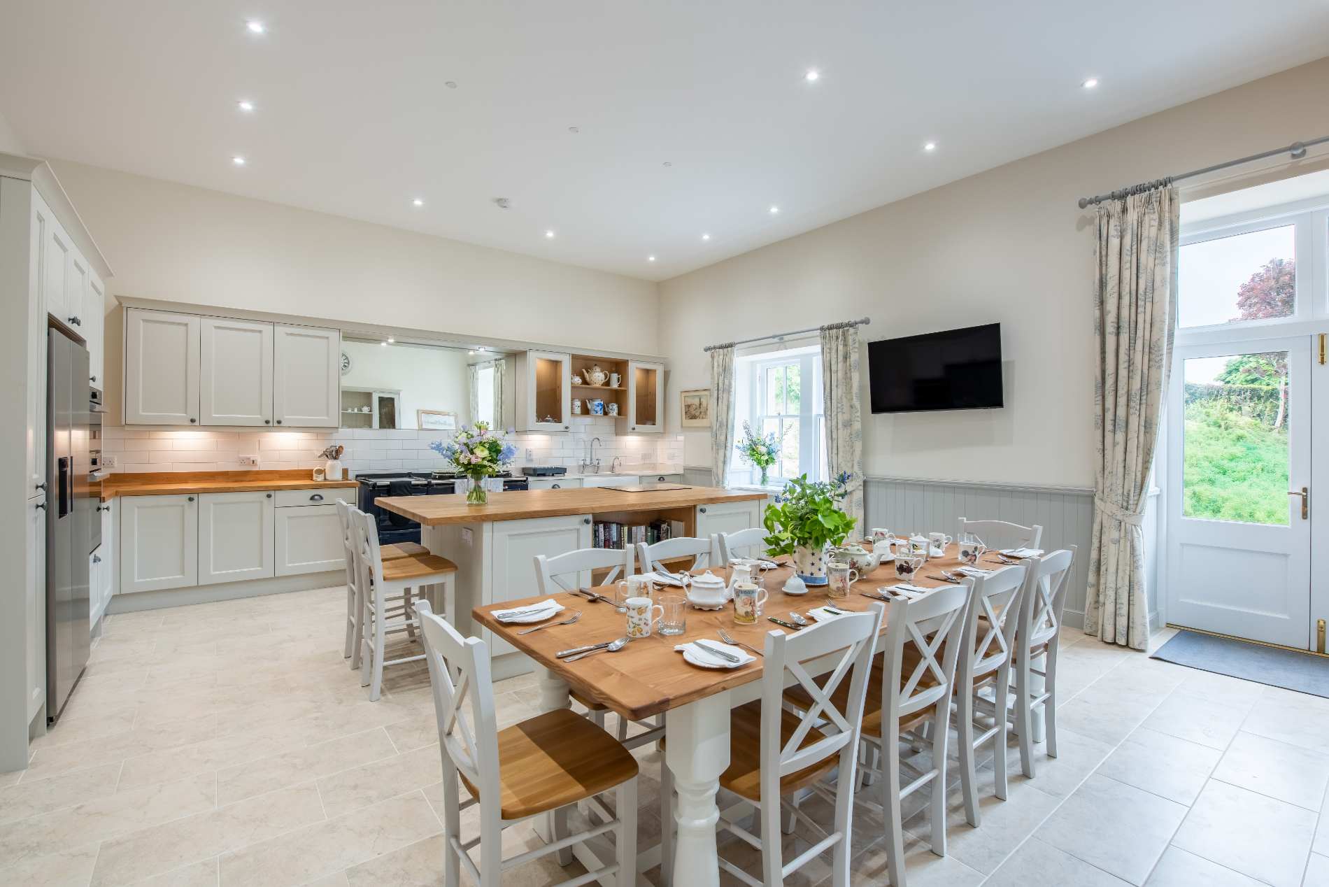A long wooden dining table is set for a group before white cabinetry, French doors opening onto the garden beyond.