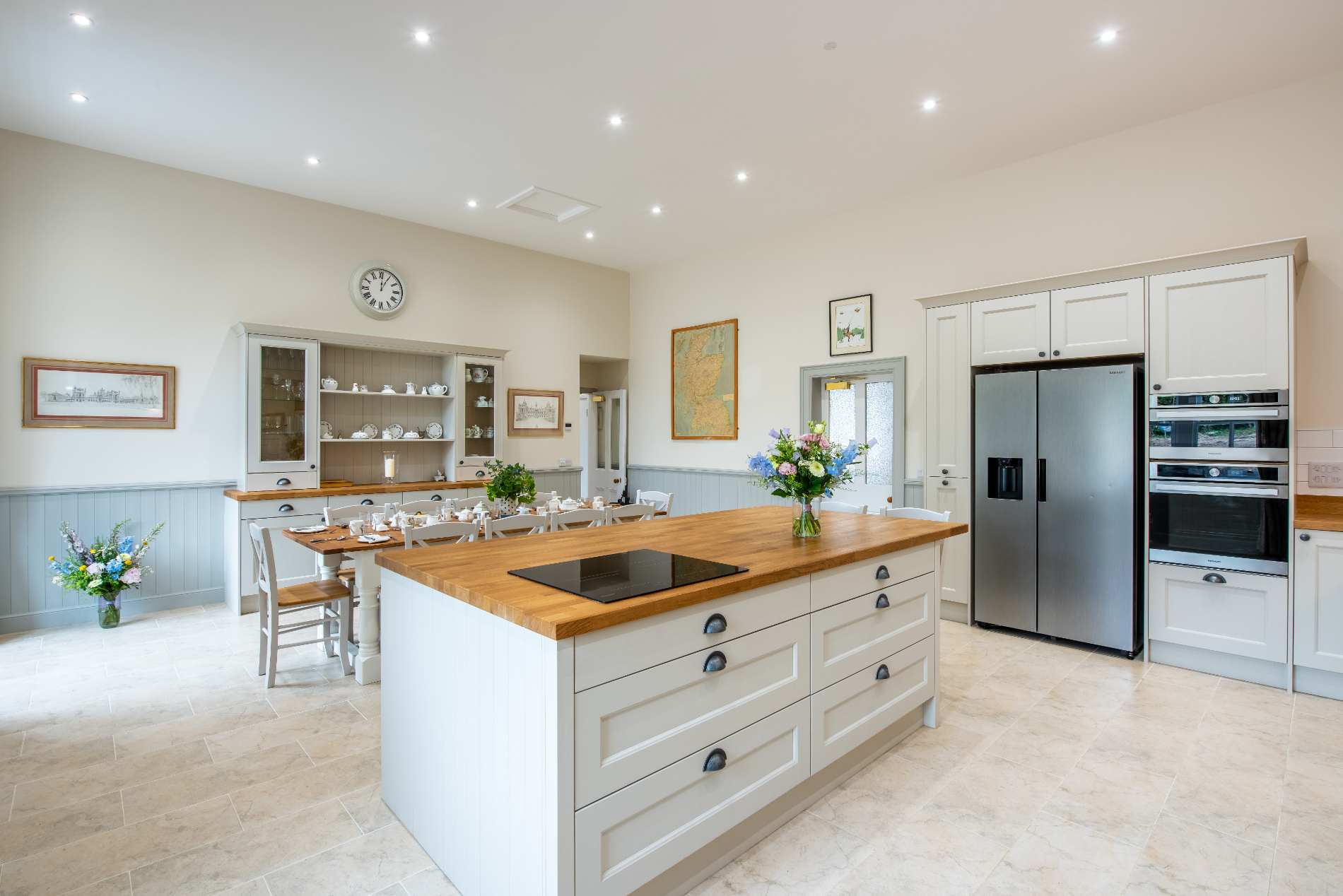 A central kitchen island with an induction hob stands before a dresser of crockery, a stainless fridge-freezer and double oven nearby a dining table.