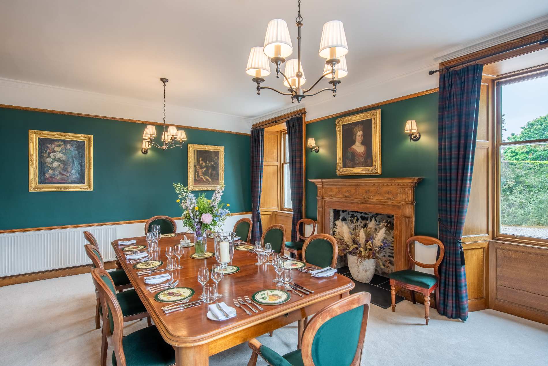 A long table is set for a group beneath a chandelier in a dark green dining room hung with gilt-framed portraits and plaid curtains.