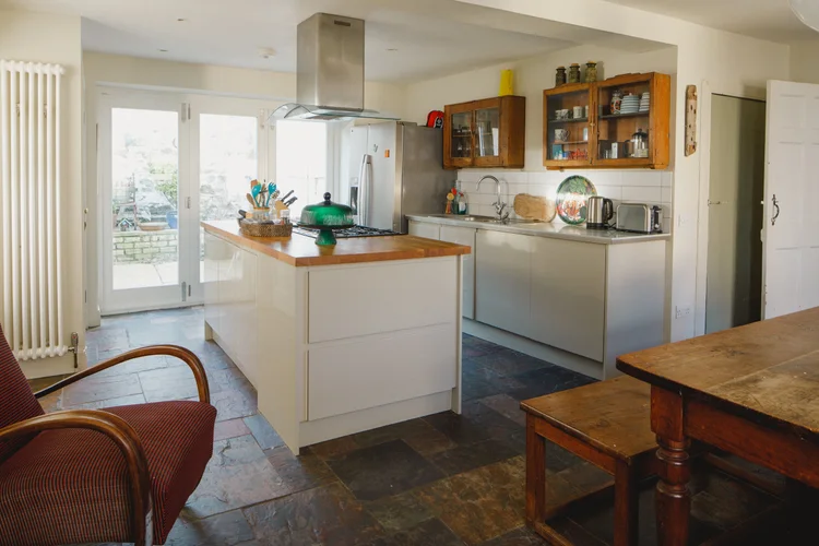 A kitchen with white cabinetry and a stainless steel extractor hood has glass-fronted cabinets of crockery and French doors leading to the garden, a dining table and bench to the right.