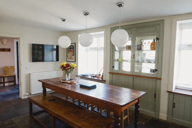 Three globe pendant lights hang above a long wooden dining table set with flowers, a glass-fronted olive cabinet and a wall-mounted television beyond.