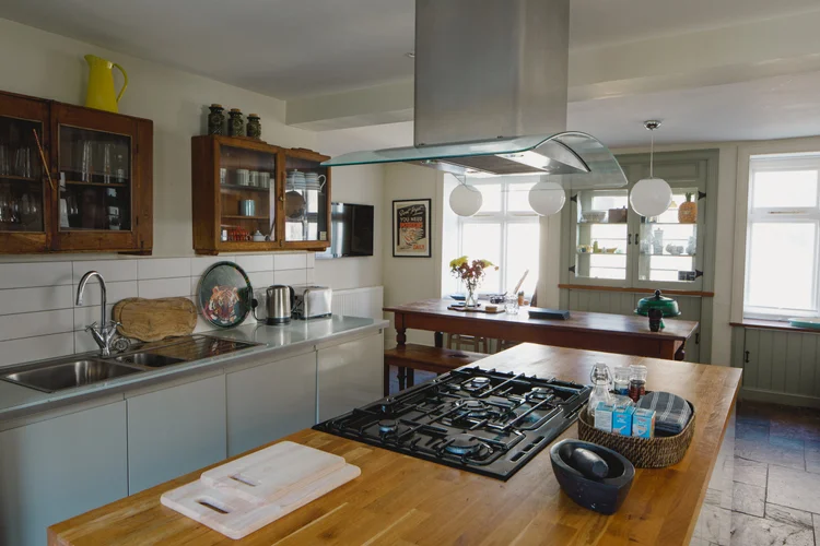 A kitchen island with a gas hob and stainless steel extractor hood stands before glass-fronted wooden cabinets, a dining table by the window beyond.