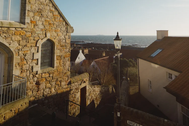 A stone church tower and rooftops are seen from above at dusk, with the sea and a lit lamppost beyond.