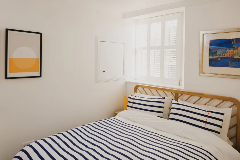 A bed dressed in navy and white stripes sits beneath a framed print, a window with white plantation shutters to the side.