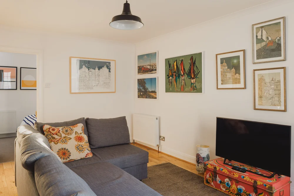 A grey corner sofa with a floral cushion sits before a gallery of framed prints, a patterned suitcase used as a stand for the television.