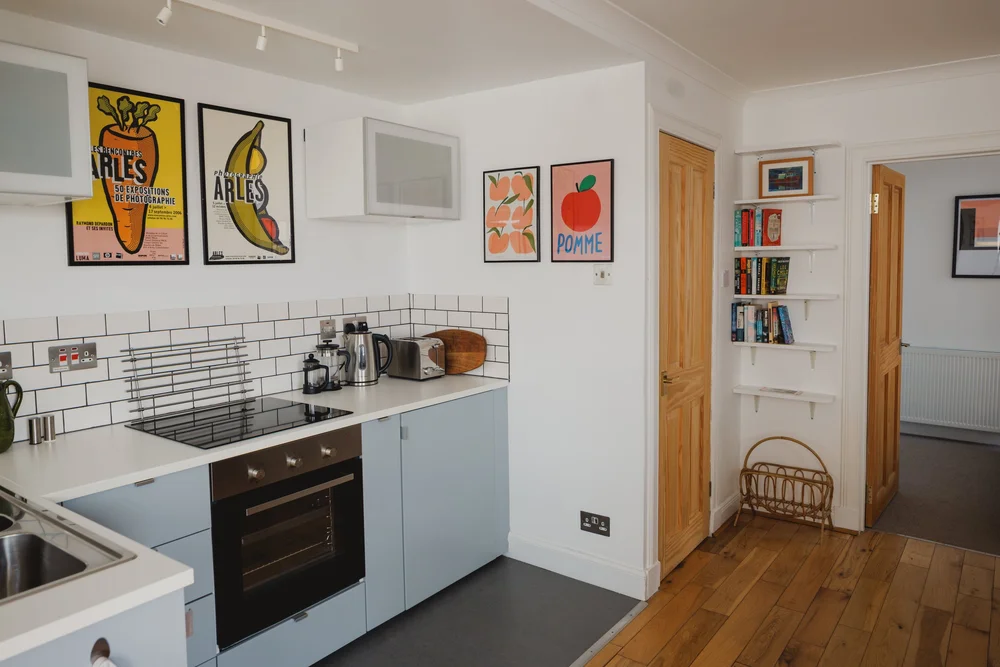 A blue kitchen with white worktops stands beneath framed fruit-themed prints, a hallway and bookshelf visible through the open doorway.