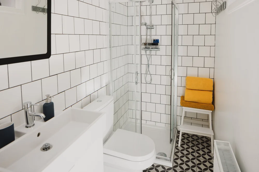A glass shower cubicle stands beside a white toilet and basin in a white-tiled bathroom, a mustard cushioned stool against the wall.