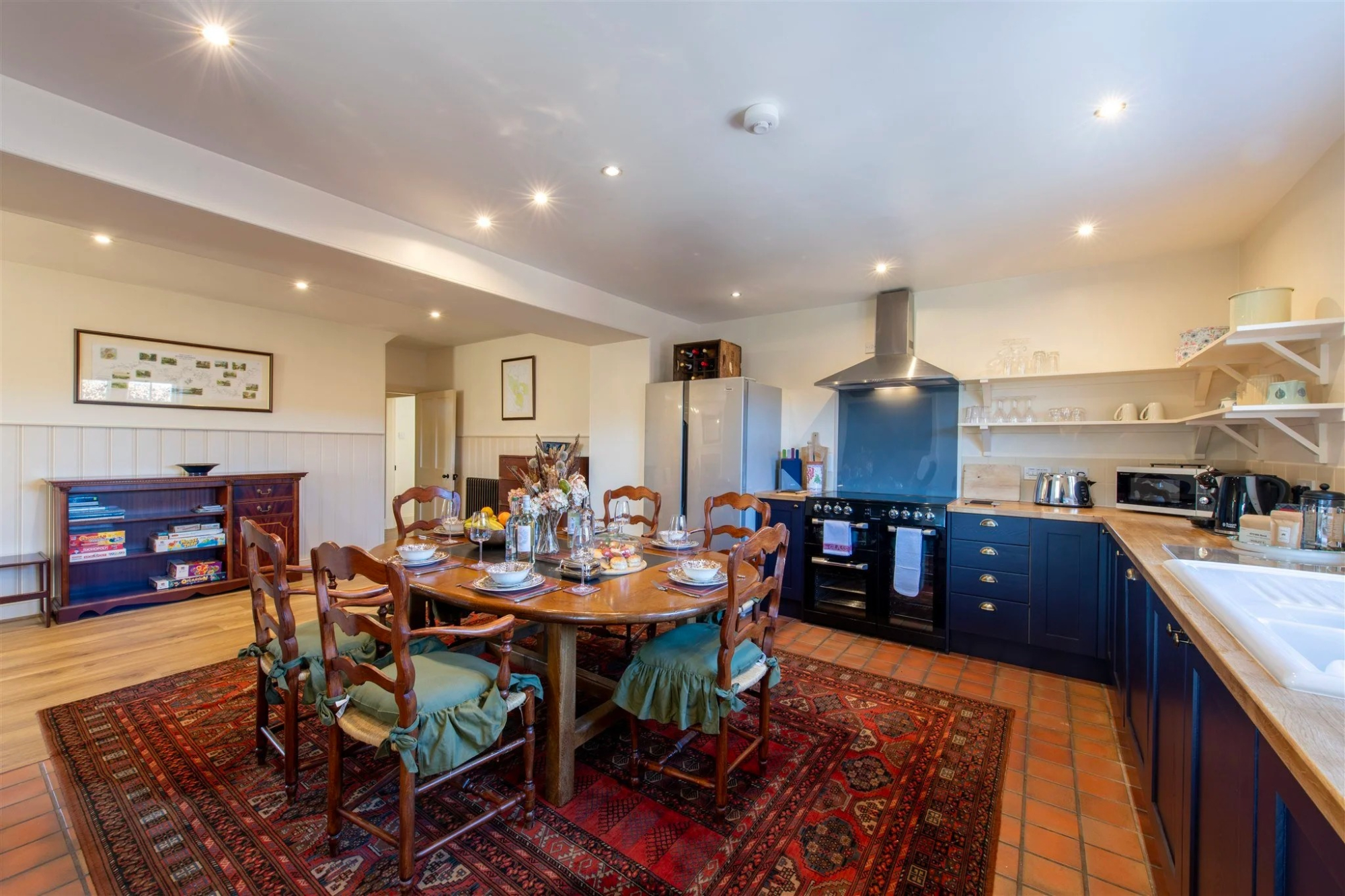 A dining table laid for a meal in front of a kitchen with dark blue cabinetry, an extractor hood and open shelving.