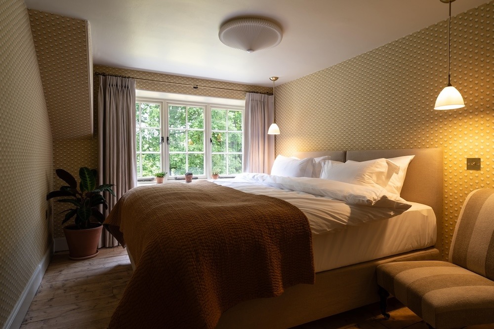 A bed with a brown throw sits before a curtained window looking onto trees, a potted plant and armchair to the side.