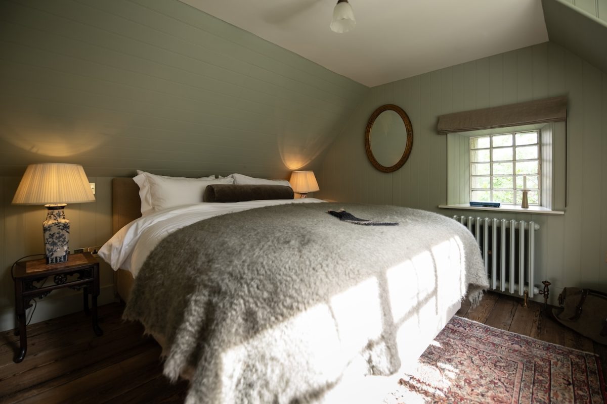 A bed with white linen sits between two lit lamps on dark wood tables, a round mirror on the sage green wall above the headboard.