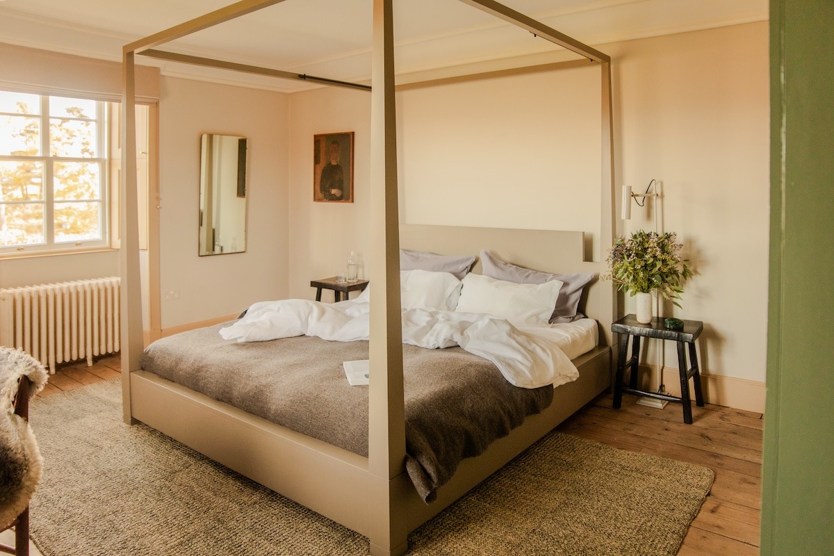 A four-poster bed frame with grey linen stands before a window, a mirror and small painting on the wall to the left.