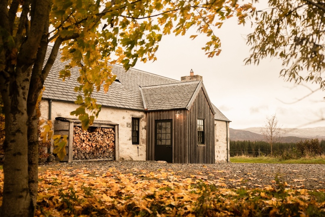 A stone and dark timber house sits beneath autumn trees, logs stacked in an open store and hills visible in the distance.