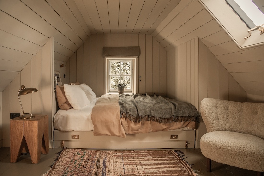 A bed with a grey throw sits in a sloped attic room, a cream bouclé chair and a wooden stool with a lamp to the sides.
