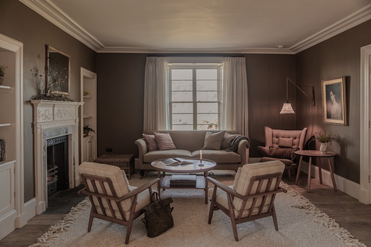 A marble fireplace sits between wingback chairs and a beige sofa in a sitting room, a pink armchair and floor lamp completing the arrangement around a round coffee table.