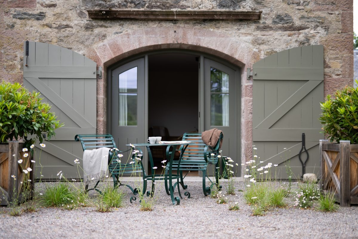 Green stable doors stand open onto a stone archway, wrought iron chairs and a table set on the gravel patio outside.