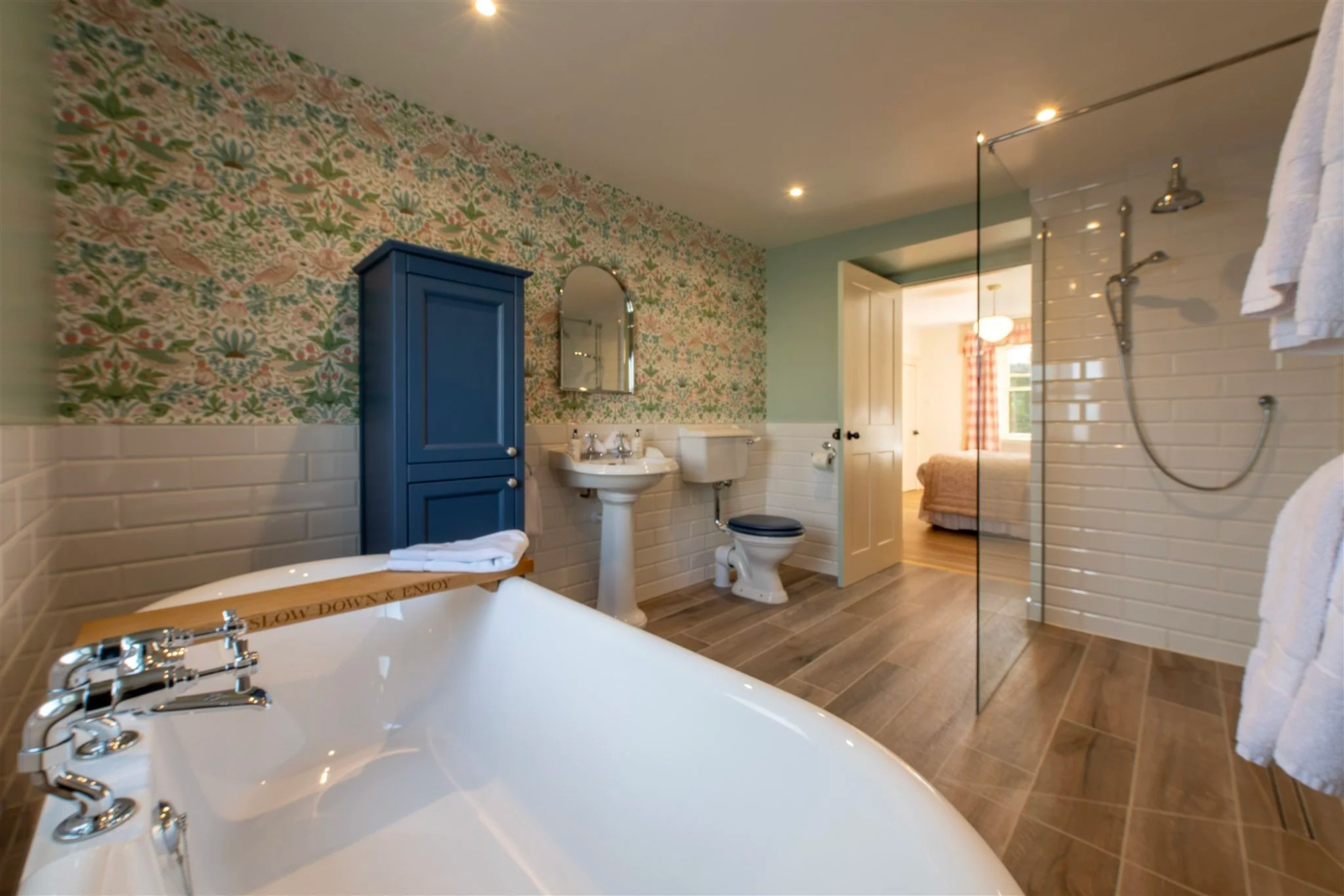 A freestanding bath faces a walk-in shower and pedestal basin in a bathroom papered with a pale floral print, a bedroom visible through the open door.